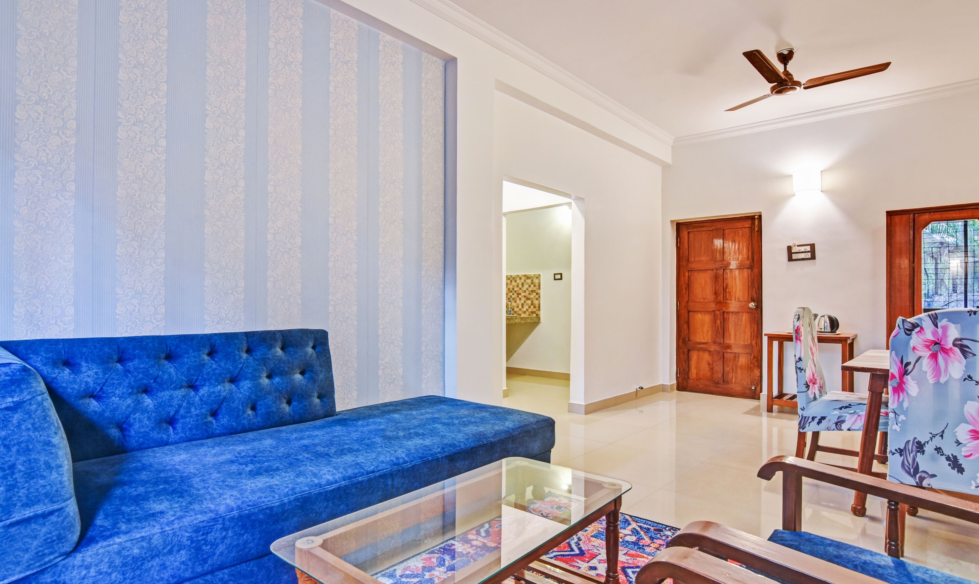 A living room with a blue sofa, a glass coffee table, and wooden chairs with floral upholstery.