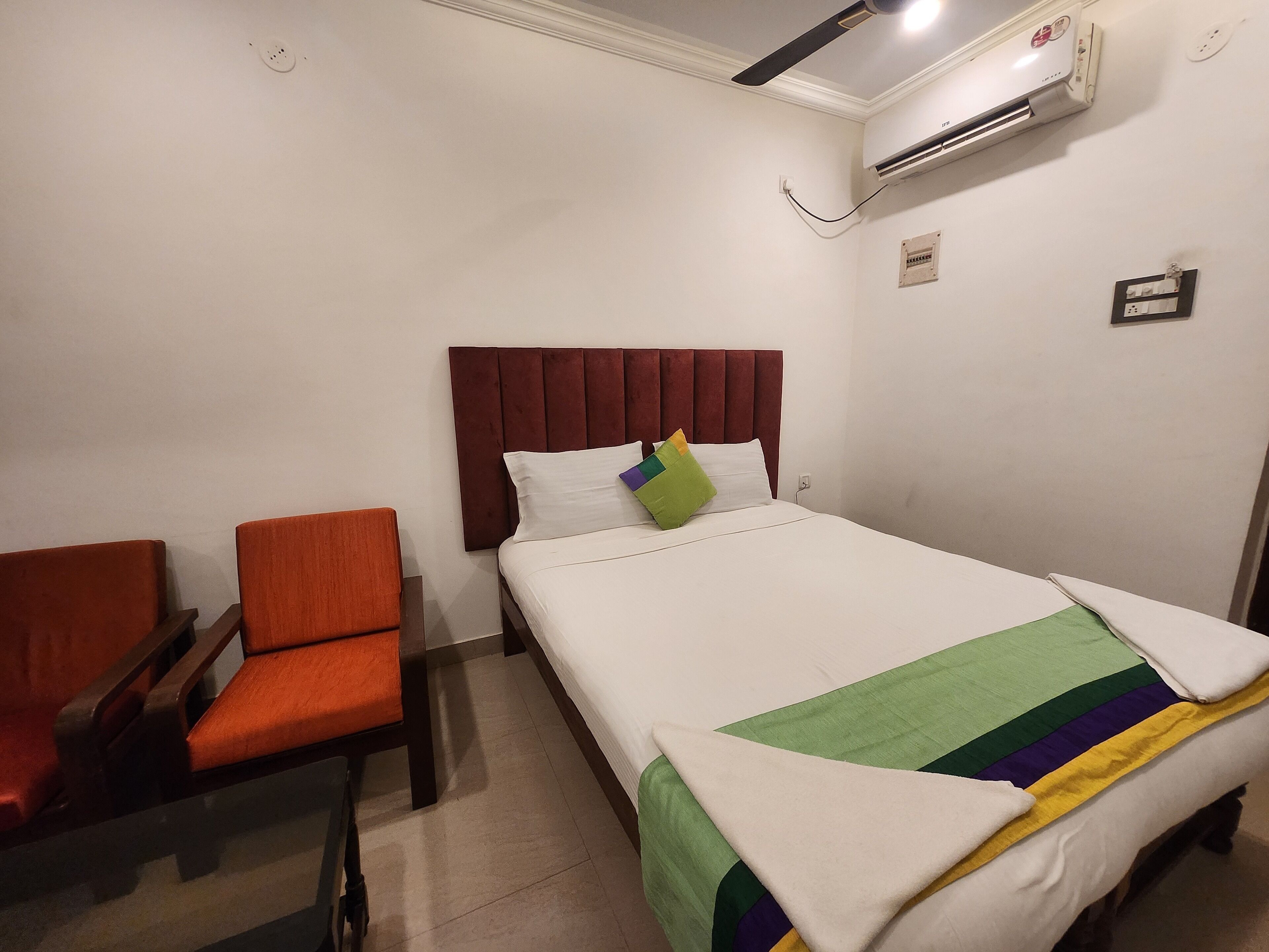 A hotel room with a bed, a chair, an air conditioning unit, and a wall socket.