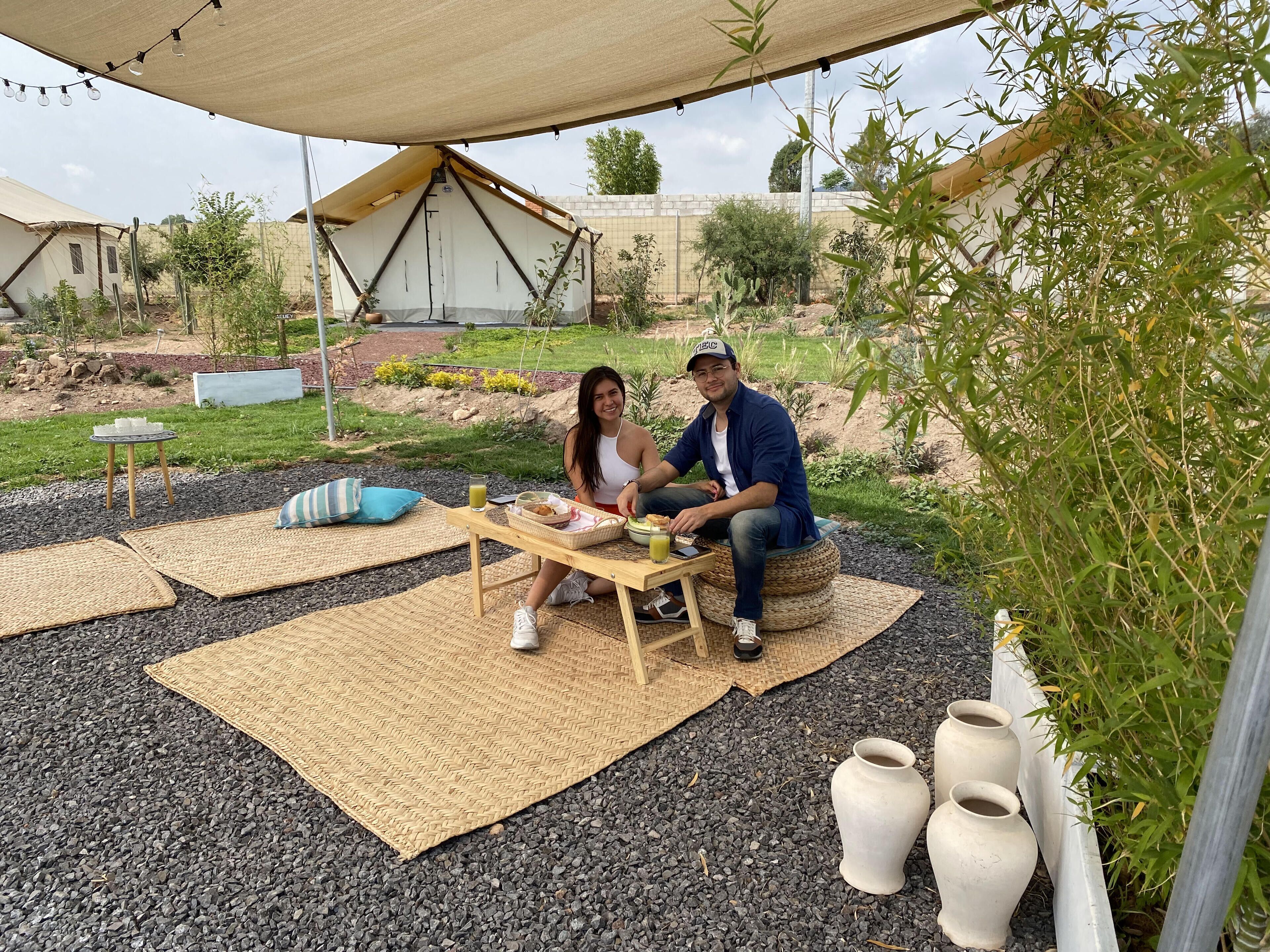Um casal sentado em mesa ao ar livre, com uma tenda ao fundo, em um jardim com plantas e um caminho de cascalho.