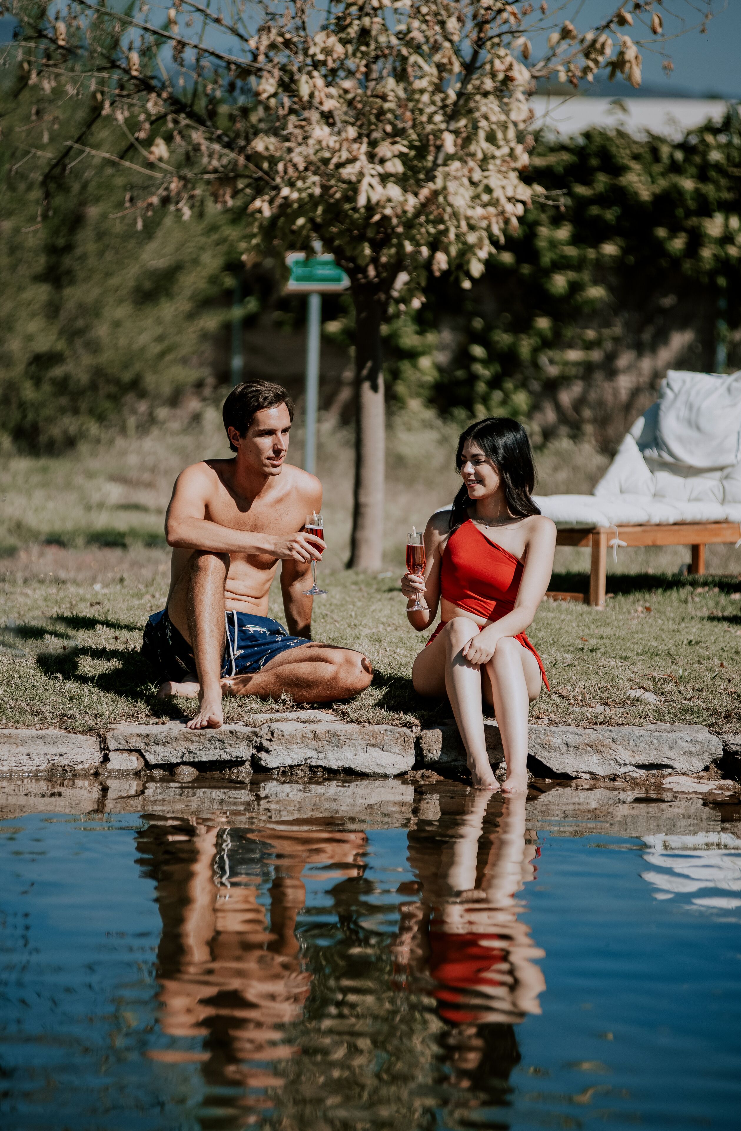 Two people sitting by a pool with a lounge chair in the background.