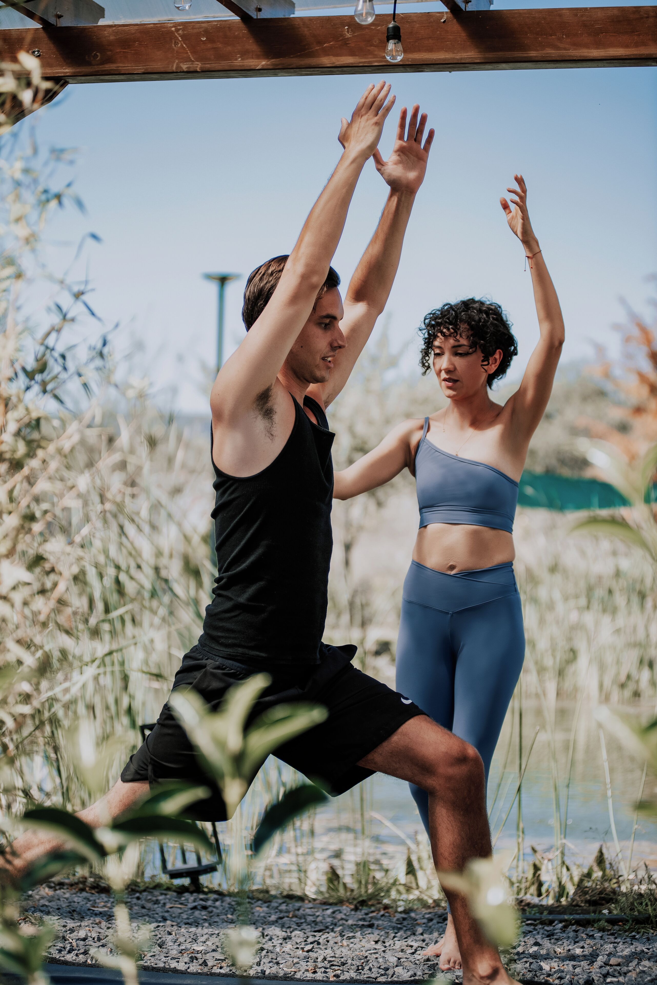 Twee mensen doen een yoga-houding buiten, met een houten constructie erboven en een heldere lucht op de achtergrond.