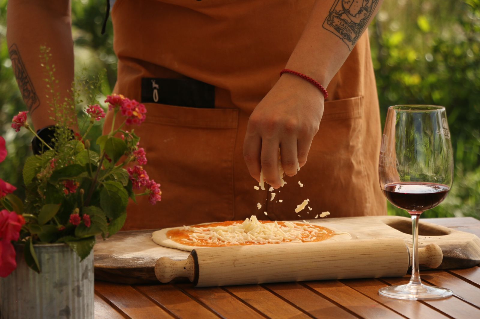 A person preparing pizza on a wooden table with a rolling pin and a glass of red wine.