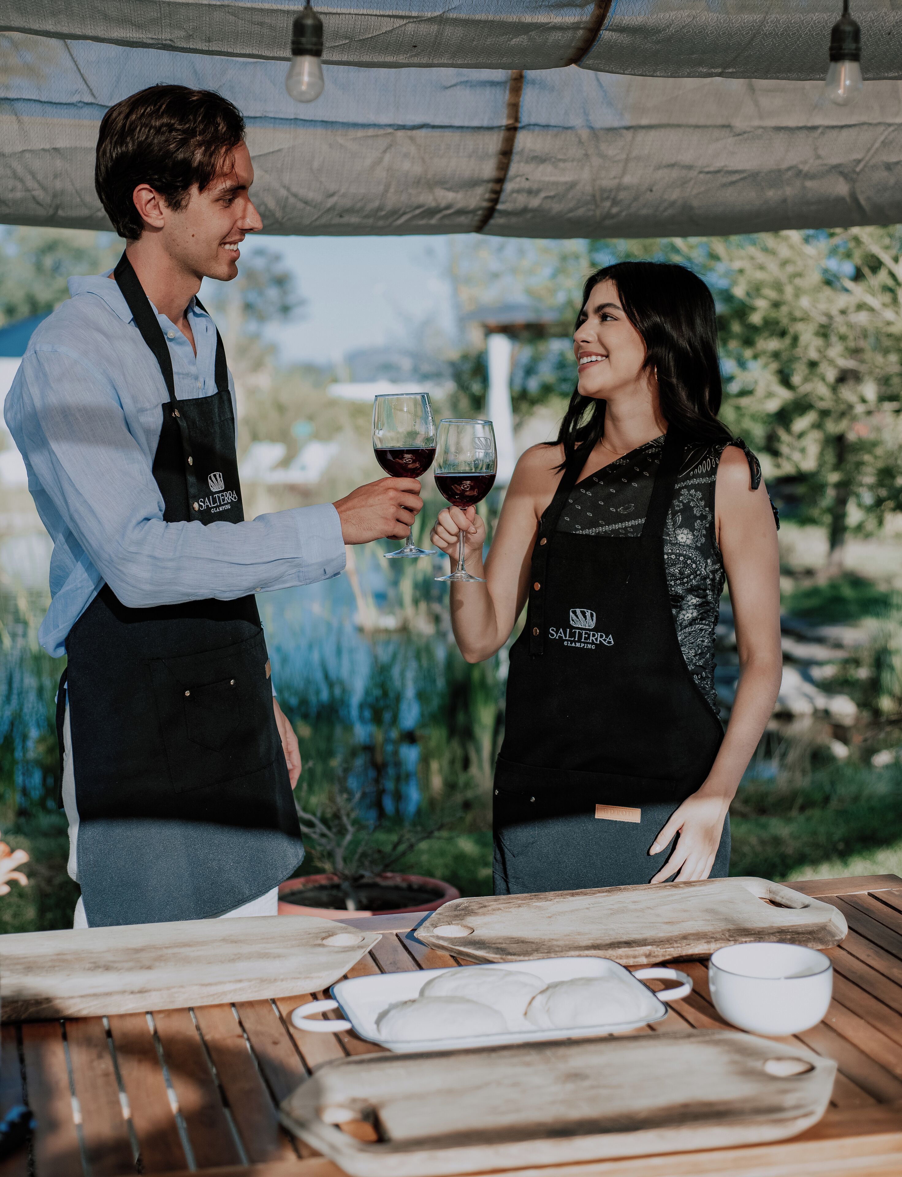 Two people toasting with wine glasses at an outdoor table with a wooden surface, surrounded by greenery.