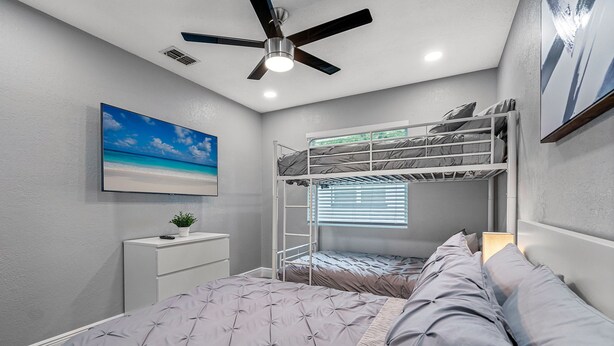 Een slaapkamer met een stapelbed, een bureau, een plafondventilator en een schilderij van een strand.