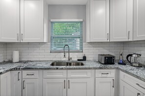 A kitchen with white cabinets, a granite countertop, a stainless steel sink, a window with blinds, and a toaster.