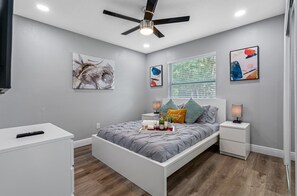 Une chambre moderne avec un lit, des tables de chevet, un ventilateur de plafond et des œuvres d’art accrochées aux murs.