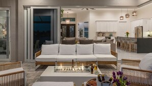 Un salon moderne avec un canapé blanc, une table basse en verre dotée d’un foyer intégré, et un coin cuisine en arrière-plan.