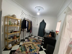 A room with a checkered floor, a wooden shelf with kitchenware, a clothing rack, a desk with a computer, and a decorative ceiling light.