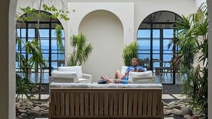 Een rustig buitenterrein met een houten bankje, omgeven door tropische planten en uitzicht op zee door grote ramen.