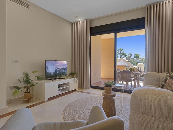 A modern living room with a flat-screen TV, comfortable seating, a dining area, and a large sliding glass door leading to a balcony with a view of palm trees.