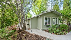 A small, single-story house with a front porch, a window with a flower box, and a garden area with shrubs and a tree. - Munson Mountain Estates (Penticton)