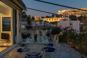An outdoor patio with a glass table and white chairs, overlooking a historic building with ruins and a hill in the background.