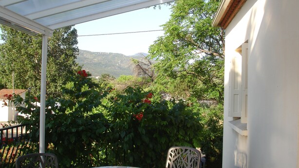 Eine überdachte Terrasse mit Tisch und Stühlen, umgeben von üppiger Vegetation und Blick auf ferne Hügel.