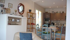 Ein Wohnzimmer mit einem beigen Sessel, einem Esstisch mit Stühlen, einer Küchenzeile mit Schränken und Geräten sowie einem Holzregal mit verschiedenen Gegenständen.