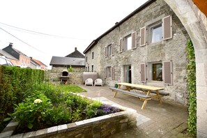 Un bâtiment en pierre doté d’un patio comprenant une table et des chaises en bois, un barbecue et un jardin avec différentes espèces de plantes.