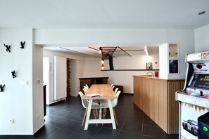 Un espace repas moderne avec une table en bois et des chaises blanches, un coin cuisine équipé d’une cuisinière, et une borne d’arcade.