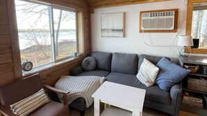 A cozy cabin interior with a wooden ceiling, a grey sofa with cushions, a white coffee table, a brown leather chair, and a window overlooking a snowy landscape. - Cabin right on the water! All bikers, skiers, fisherman, and more welcome! (Cable)