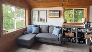 A cozy living room with a wooden ceiling, a gray sectional sofa with blue cushions, a small table with a lamp, and a wall-mounted air conditioner. - Cabin right on the water! All bikers, skiers, fisherman, and more welcome! (Cable)