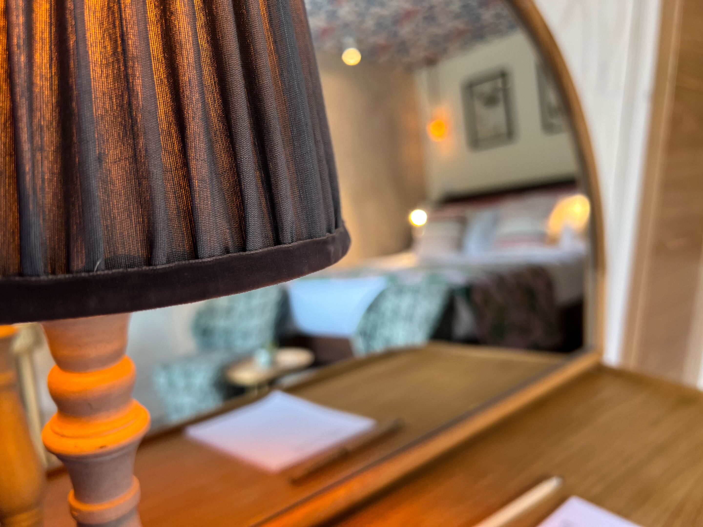 Un gros plan d’une lampe avec un abat-jour en tissu texturé, une table en bois et le reflet d’une pièce dans laquelle on aperçoit un lit et une chaise.
