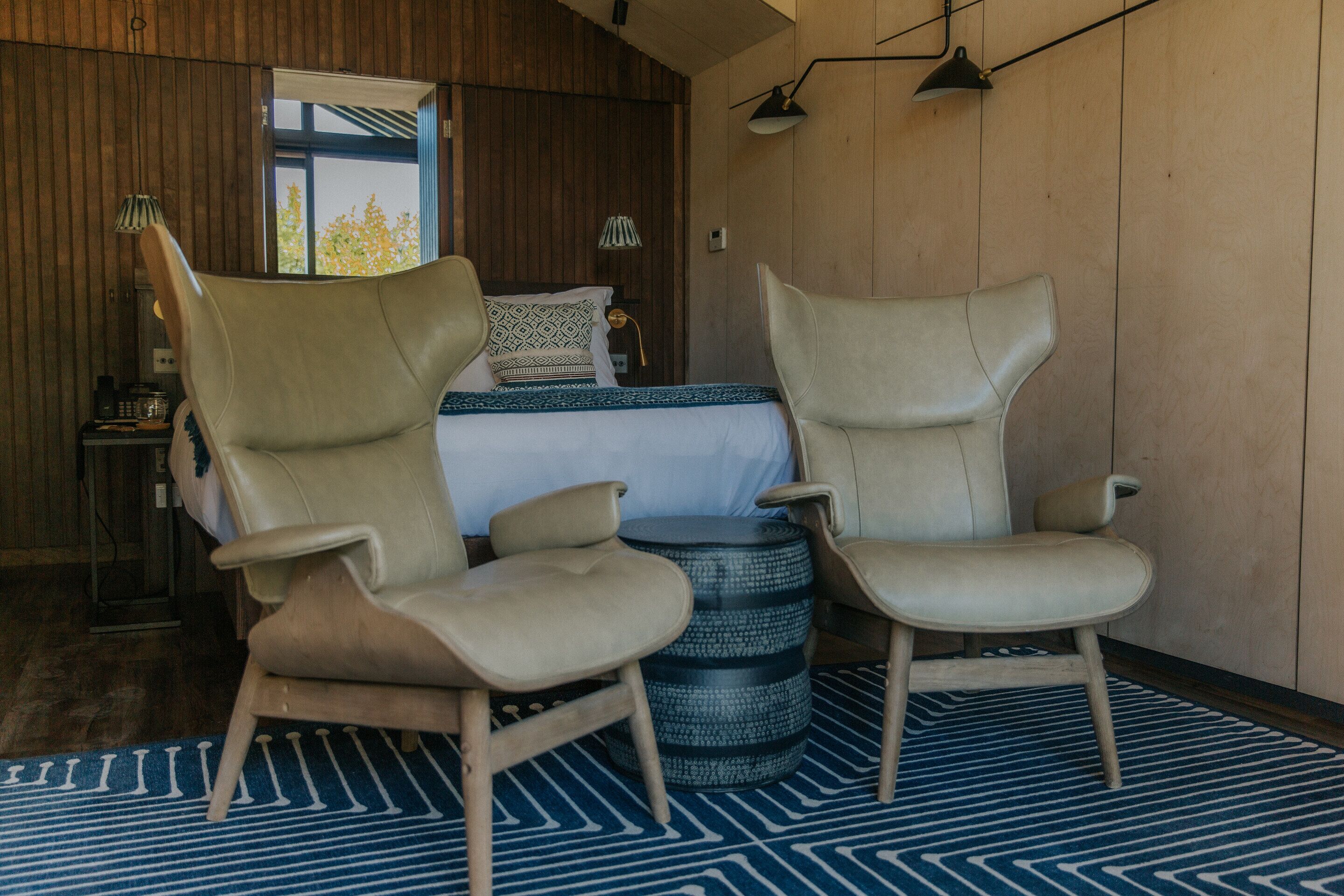 Um quarto com uma cama, duas poltronas, uma mesa redonda e um tapete estampado.