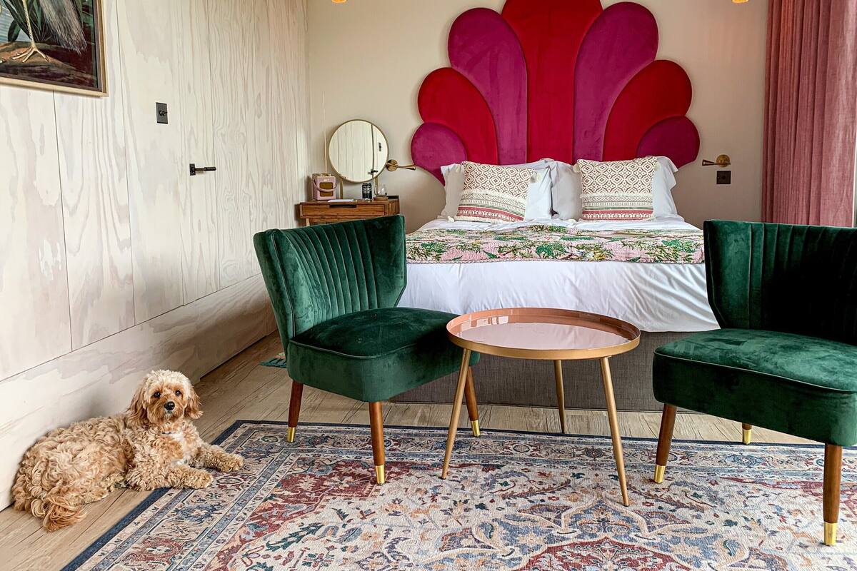 Un dormitorio con una cama grande, dos sillones de terciopelo verde, una mesita redonda, un perro y un panel decorativo en la pared.