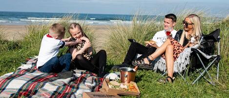 A group of people sitting on a blanket by the beach with pizza boxes and a drink.