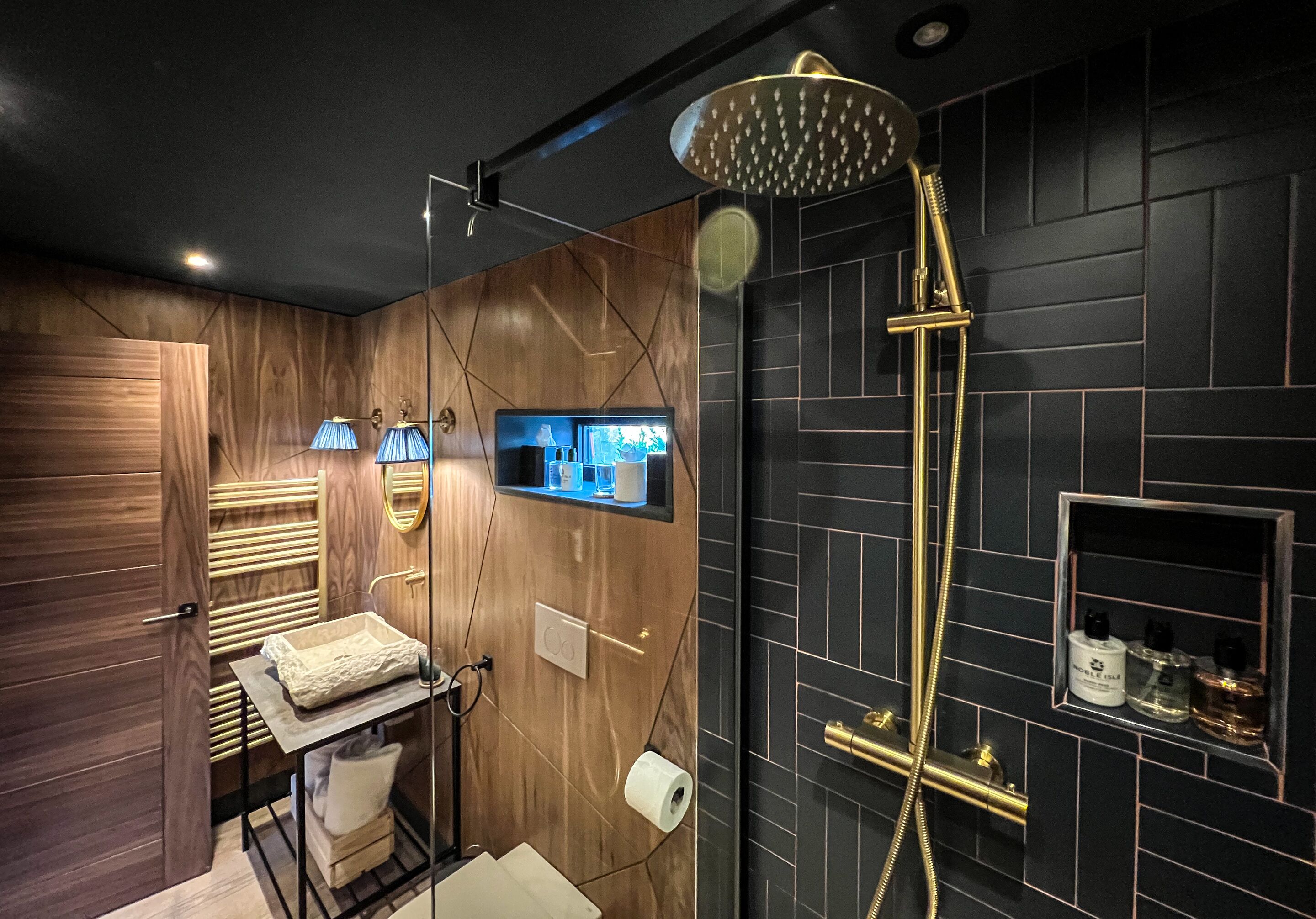 A modern bathroom with a wooden vanity, a built-in TV, a shower with a golden showerhead, and a small shelf with toiletries.