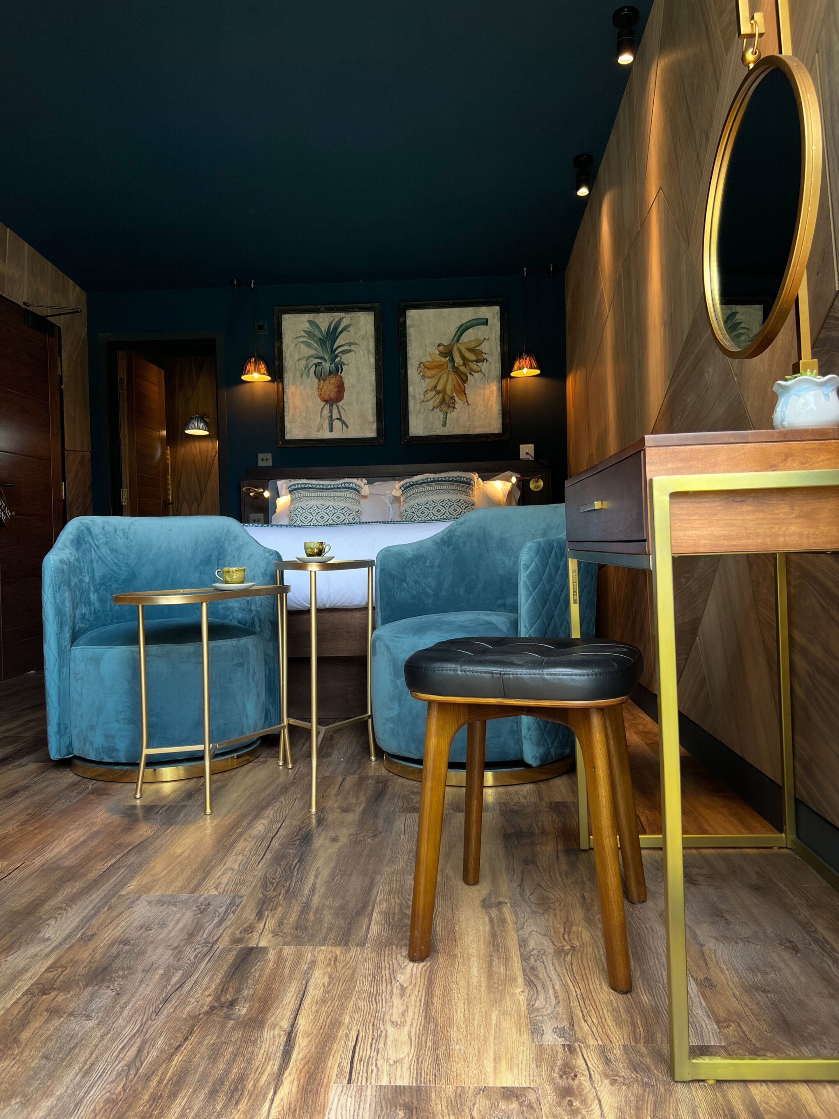 Een kamer met een bed, twee blauwe fauteuils, een klein tafeltje, een houten bureau en een spiegel.