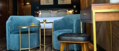 Een kamer met een bed, twee blauwe fauteuils, een klein tafeltje, een houten bureau en een spiegel.