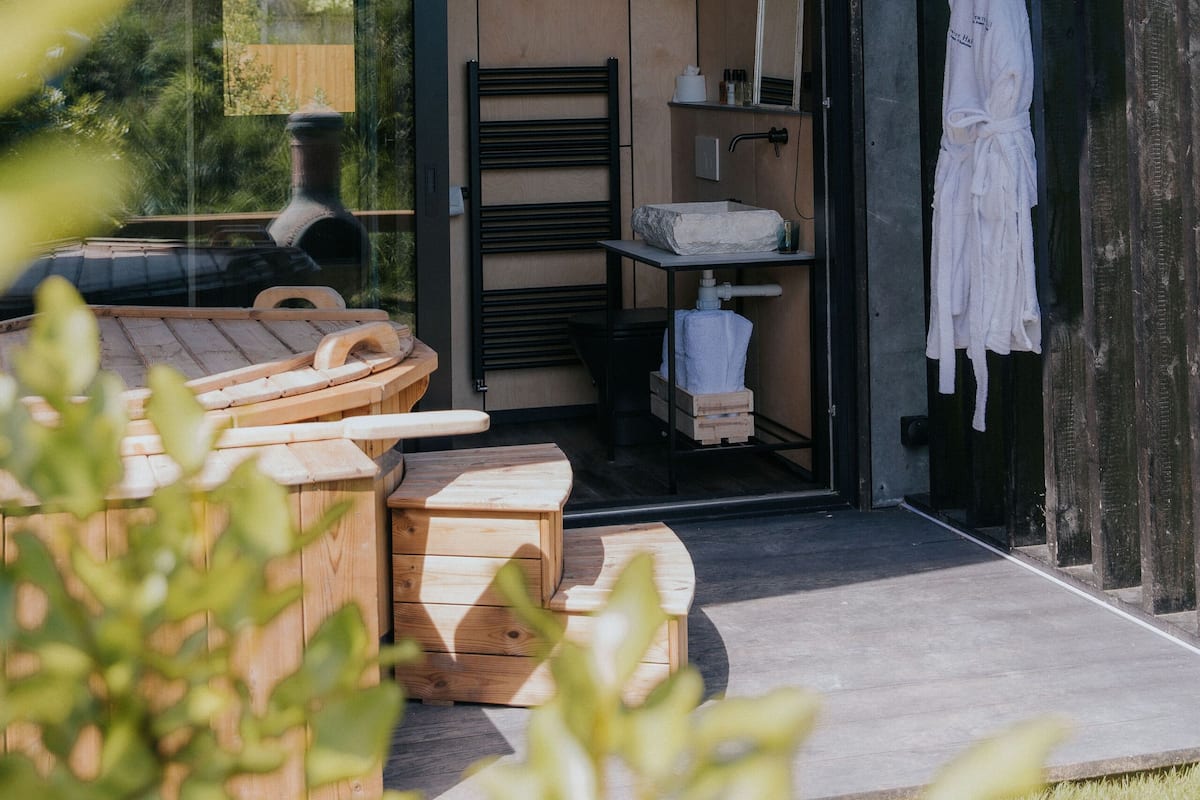 Un baño exterior moderno con puerta de vidrio, muebles de madera y una bata blanca colgada en la pared.