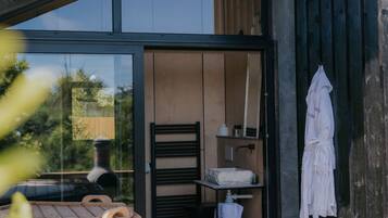 A modern outdoor bathroom with a glass door, wooden furniture, and a white robe hanging on the wall.