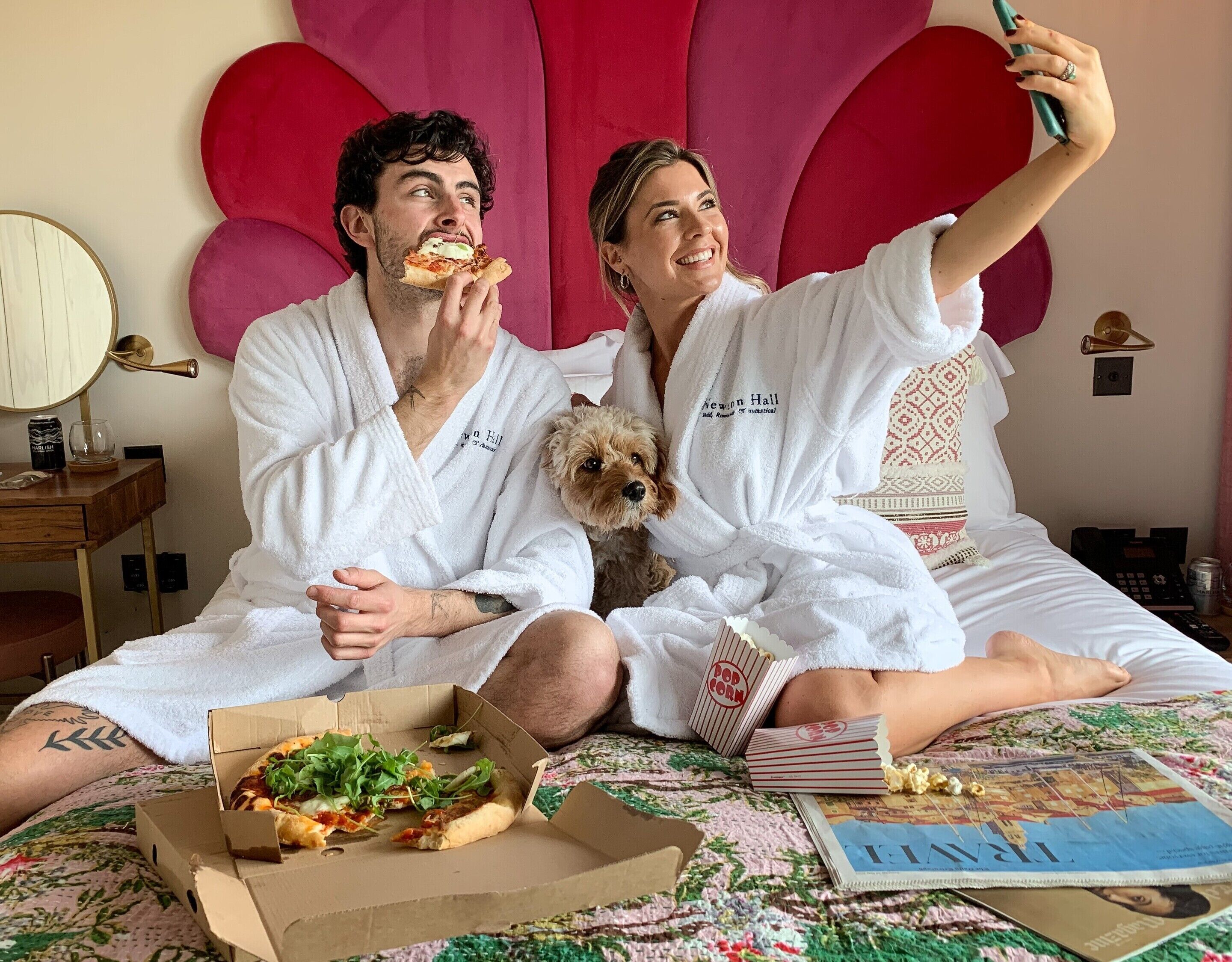 A couple in white bathrobes sitting on a bed with pizza boxes, a dog, and a magazine.