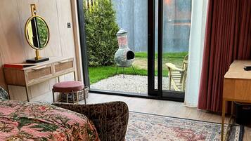 A bedroom with a bed, a small stool, a wooden nightstand, a mirror, and a view of a garden through a sliding glass door.