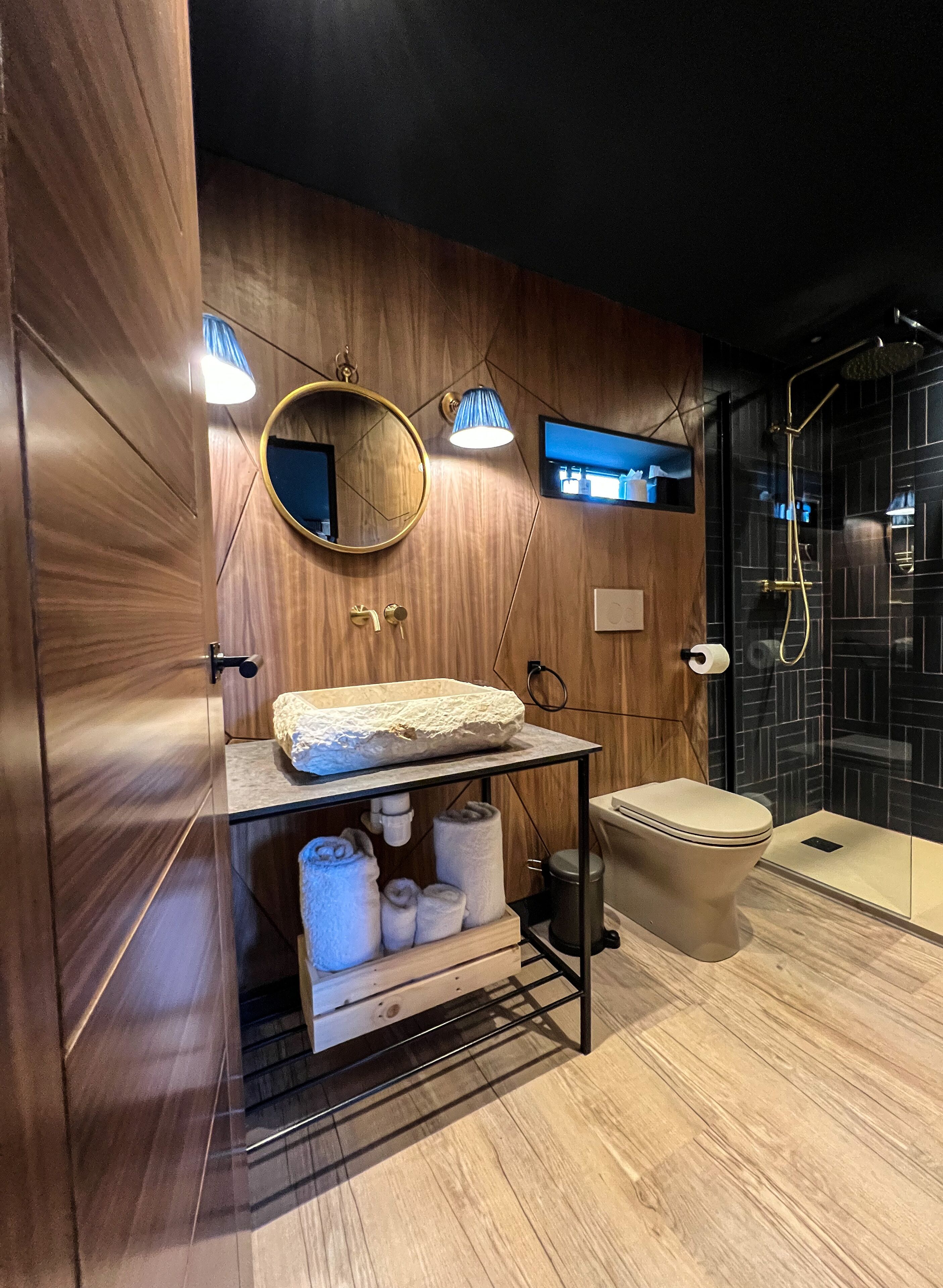 A modern bathroom with a wooden vanity, a round mirror, a toilet, and a glass-enclosed shower area.