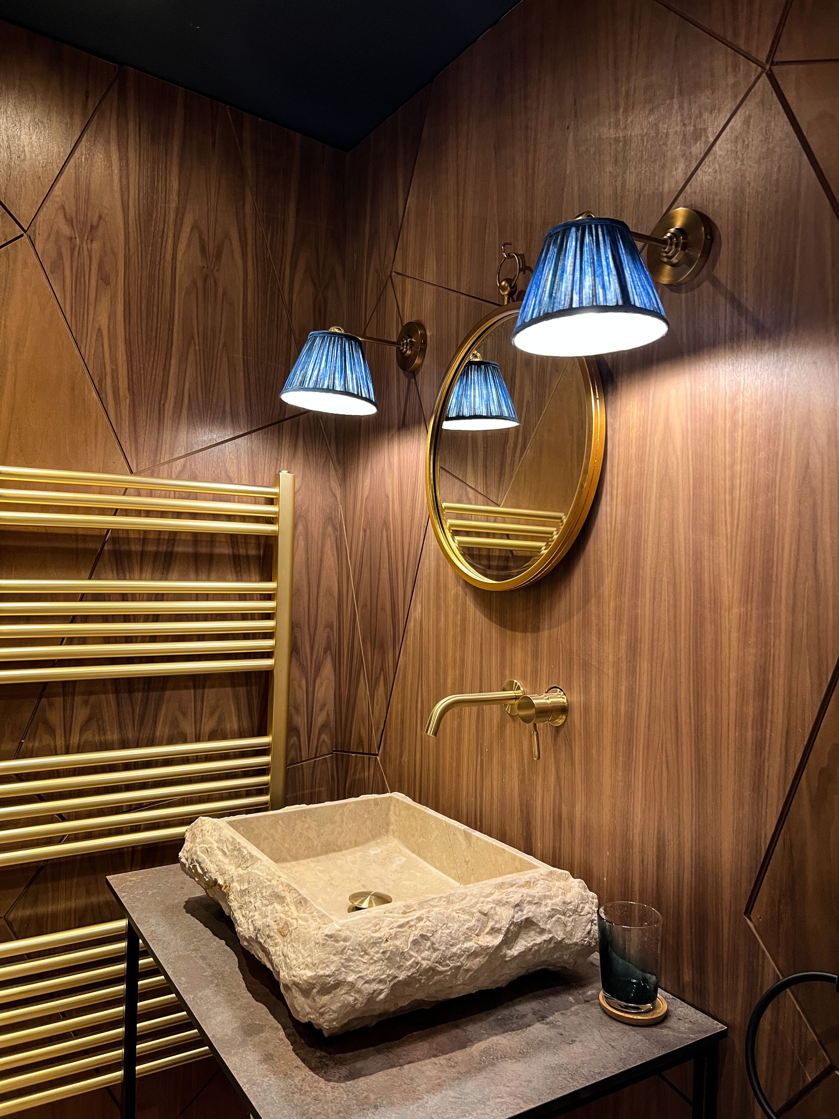 A bathroom with a wooden wall, a gold-framed mirror, a sink with a stone-like countertop, and two blue lamps.