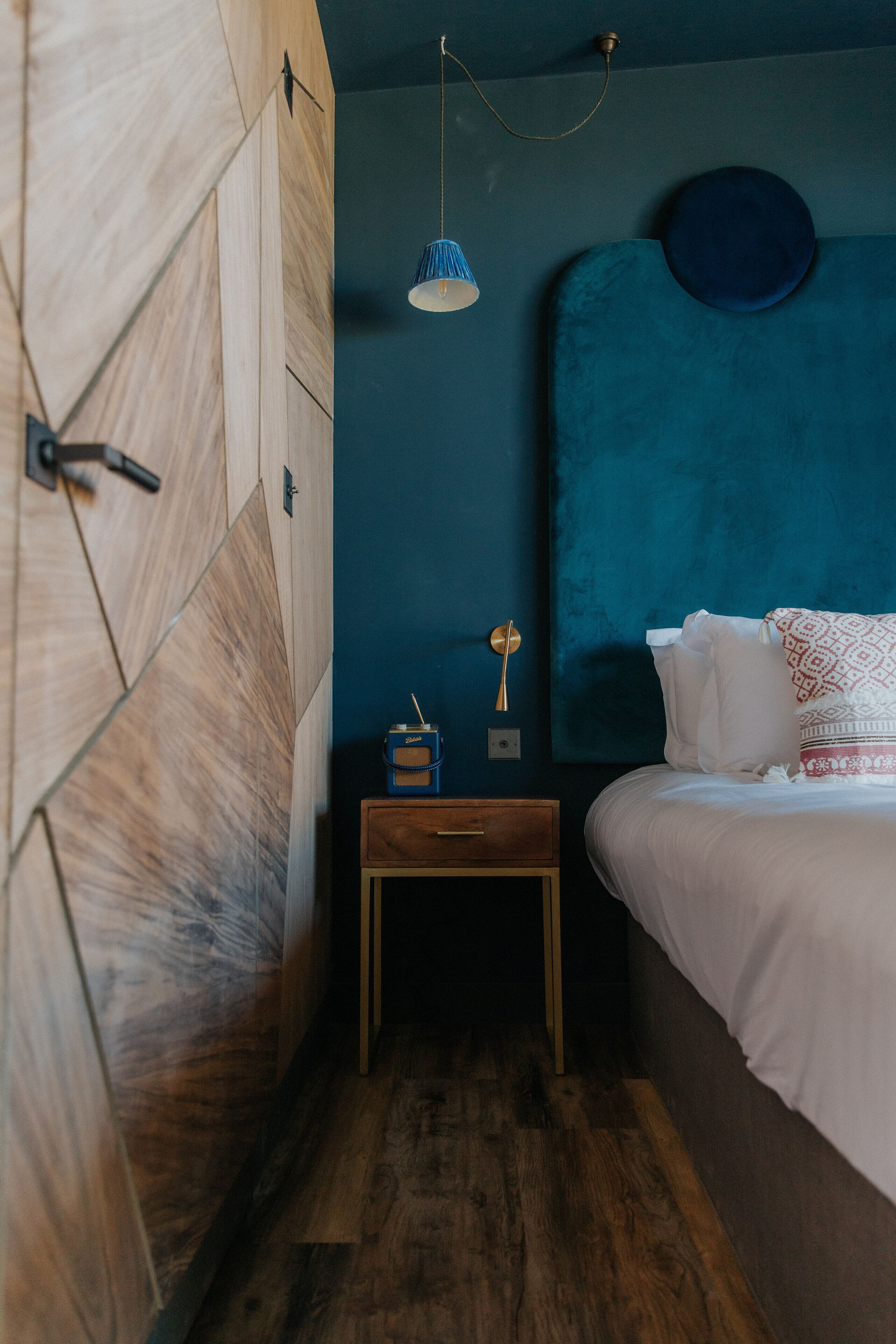 A bedroom with a wooden wardrobe, a bedside table with a lamp, and a bed with a blue headboard.