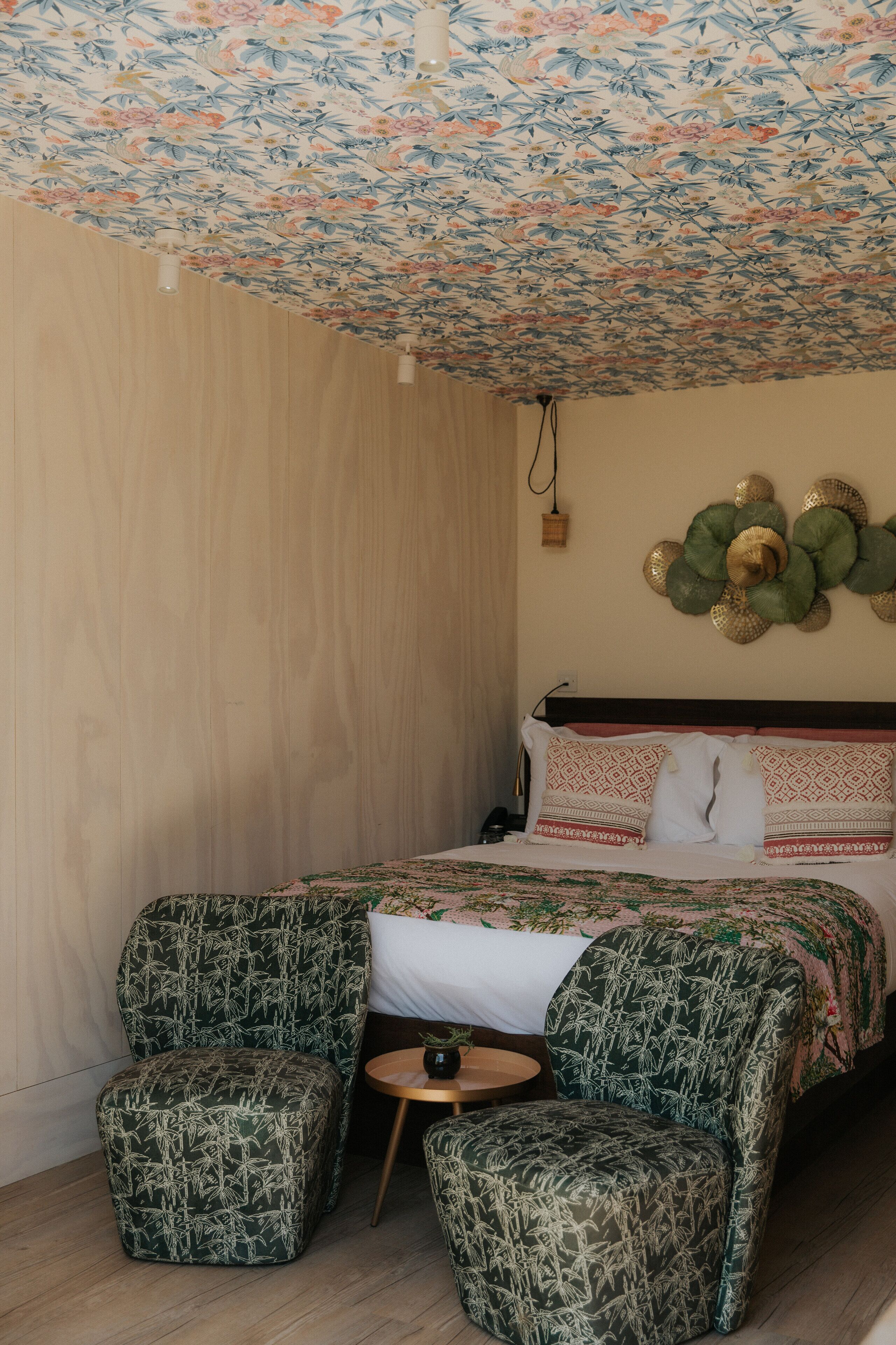 Een slaapkamer met een bloemendecor op het plafond, een bed met beddengoed met een patroon, twee bijpassende fauteuils en een kleine ronde tafel.