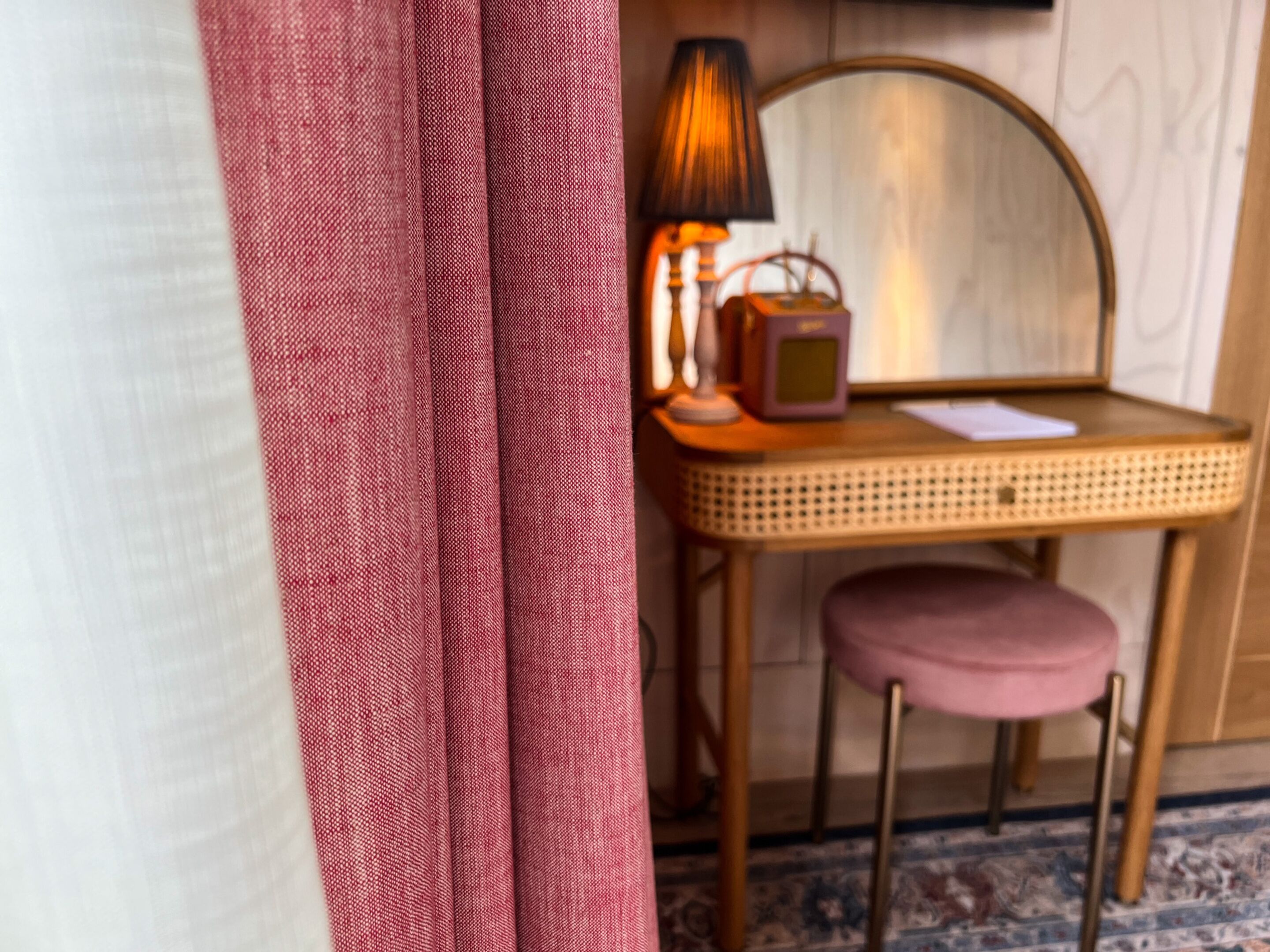 A room with a wicker chair, a small table with a lamp, and a pink cushion.
