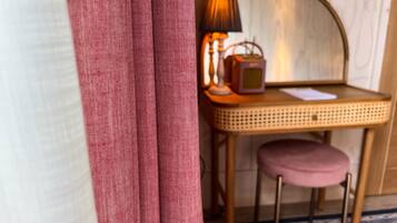 A room with a wicker chair, a small table with a lamp, and a pink cushion.
