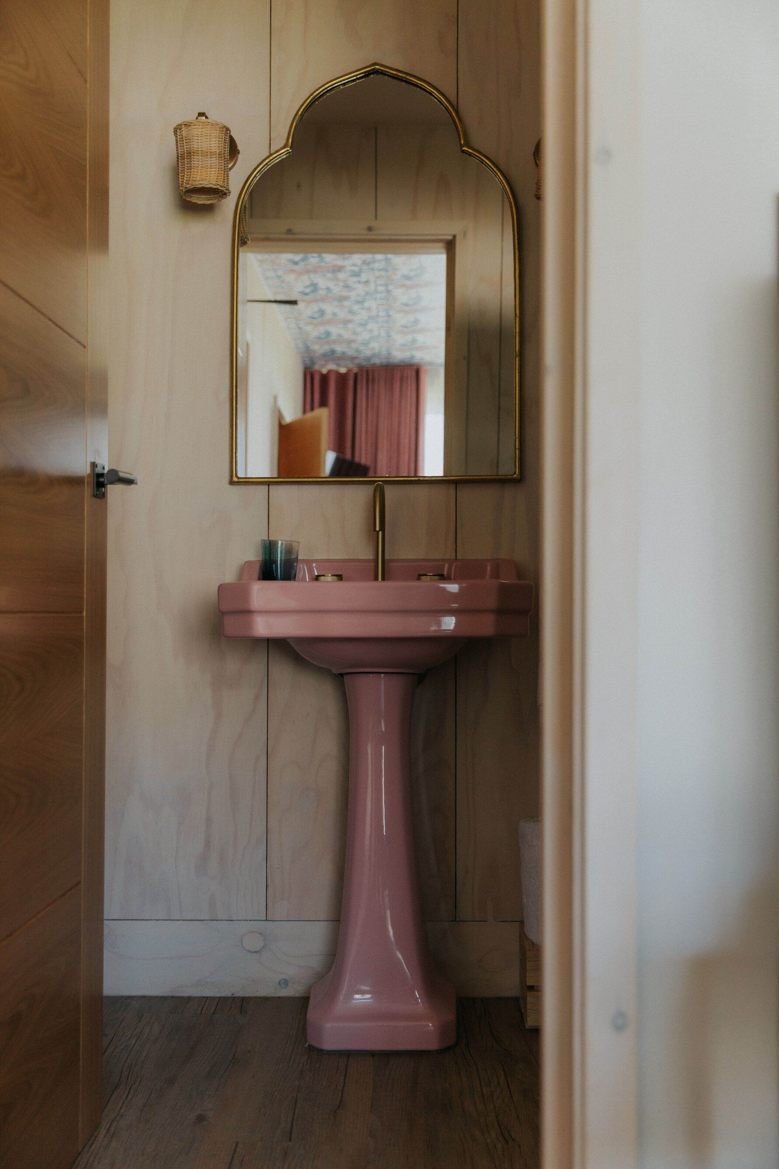 Een badkamer met een houten deur, een roze wastafel en een spiegel met gouden lijst.