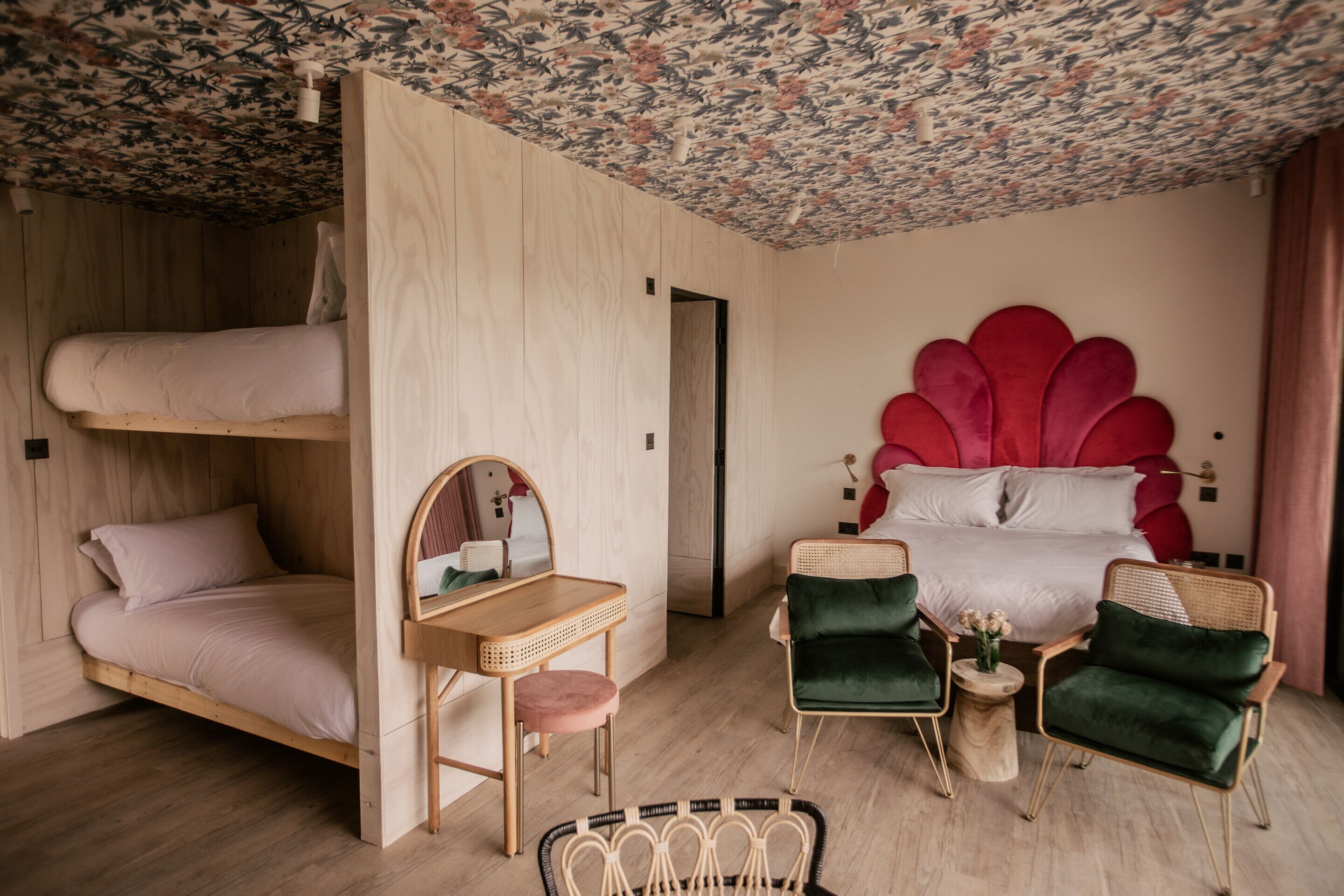 A room with a bunk bed, a desk with a mirror, two green armchairs, and a red decorative headboard.