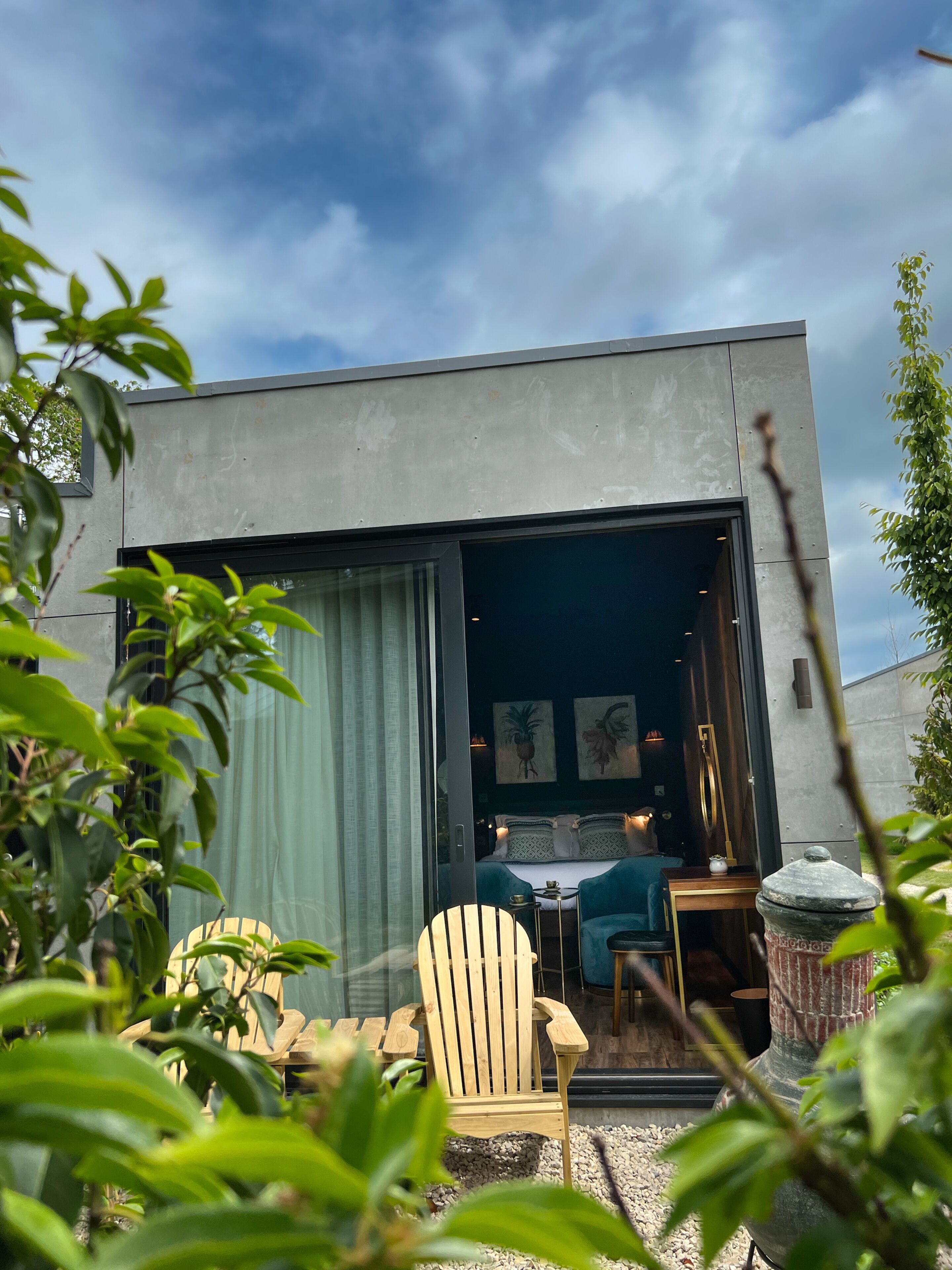 A modern building with a large sliding glass door leading to a cozy interior. Inside, there is a bed with a blue headboard, a small table with a lamp, and a chair.
