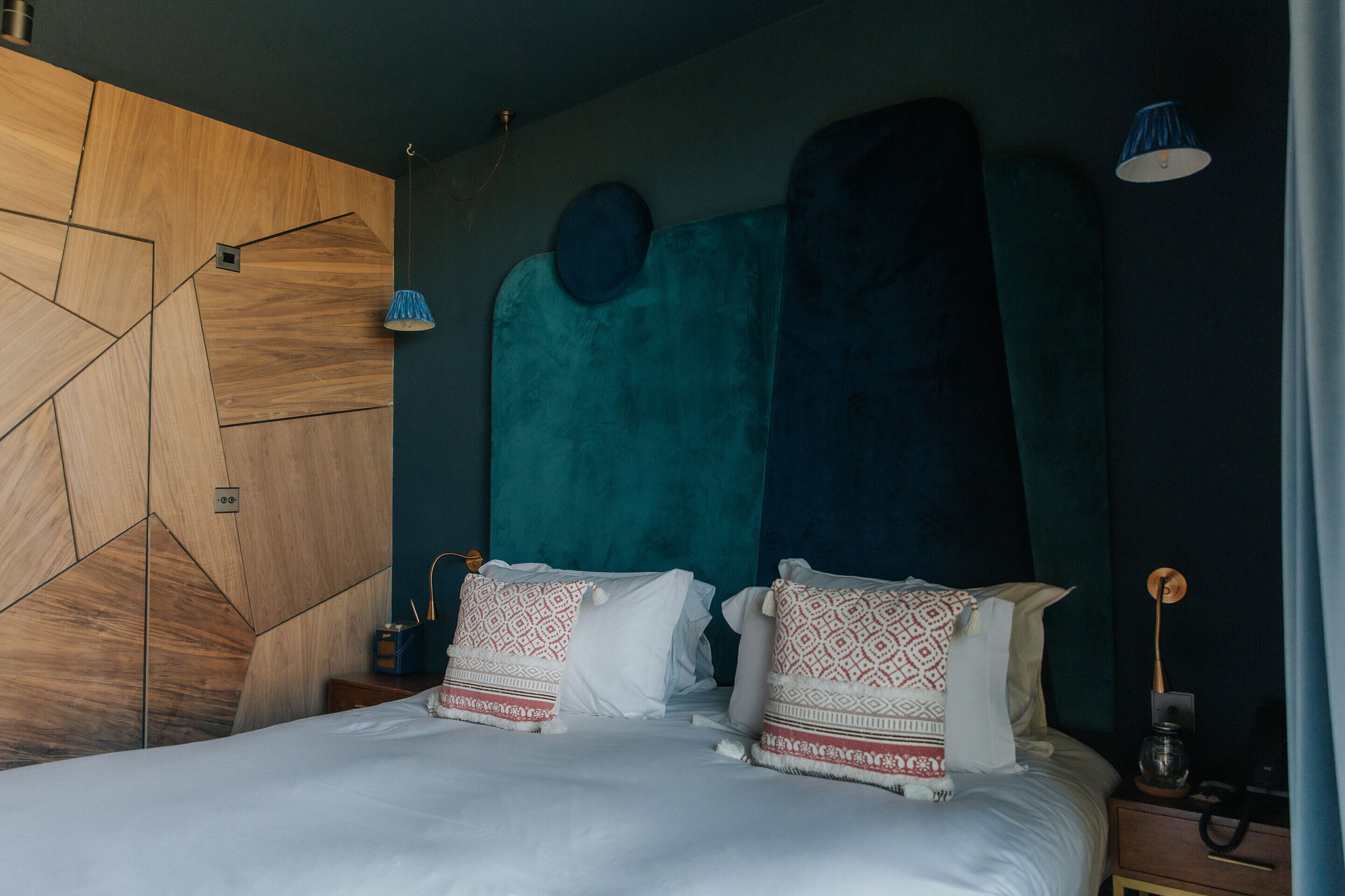 Un dormitorio moderno con un cabecero de madera con diseño geométrico, una pared de acento color aguamarina y una colcha blanca con almohadas estampadas.