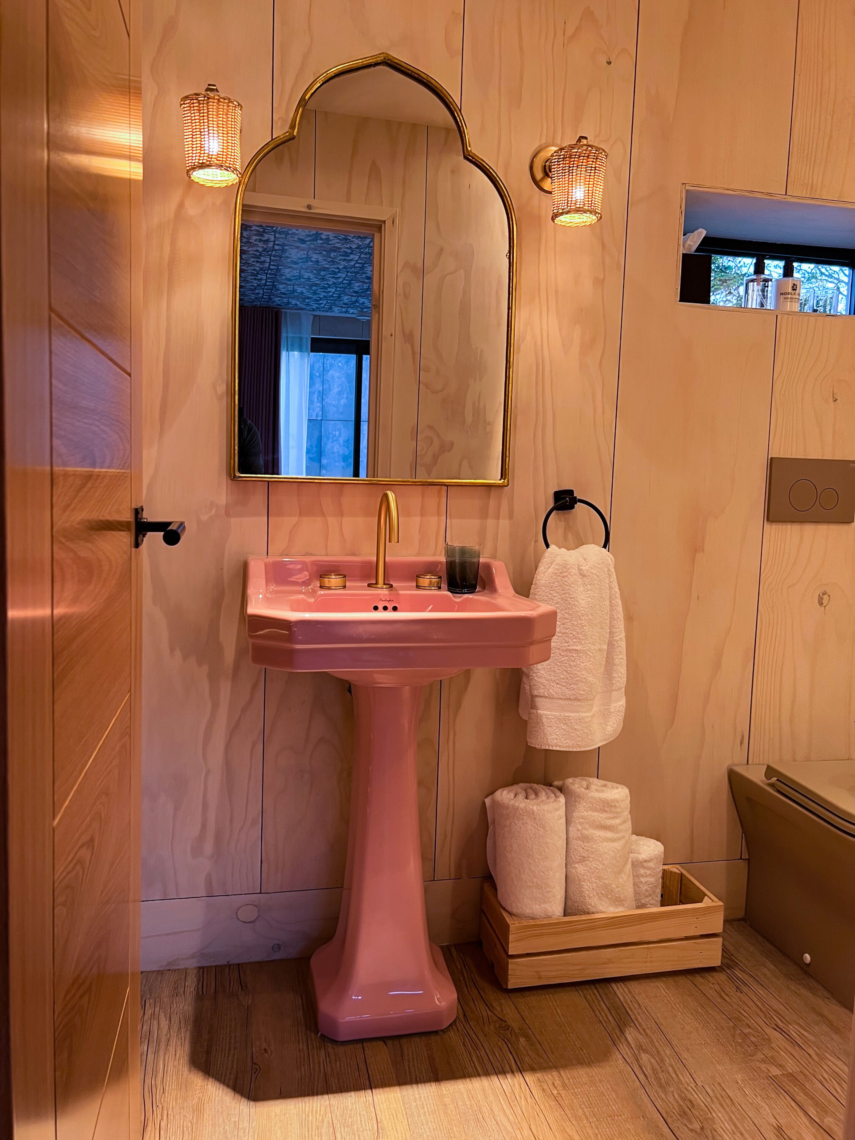 Et badeværelse med en pink vask, et spejl og trævægge. Der hænger to håndklæder på kroge, og en kurv med håndklæder i træ.