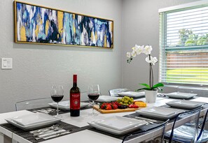 Una mesa de comedor preparada con platos, cubiertos, vasos y una botella de vino, con un cuadro en la pared y una ventana con persianas.