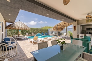 Un área junto a la piscina con sillones de descanso, una mesa de vidrio y un parasol de paja.