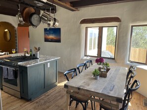 Una cucina con una grande isola, tavolo da pranzo in legno e sedie. Pentole appese sopra l'isola, un quadro al muro e una finestra con vista sugli alberi.