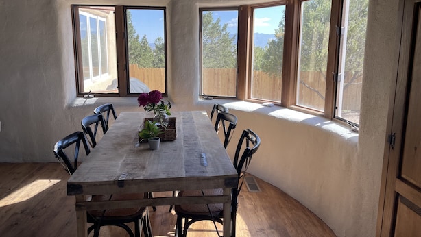 Un coin repas avec une table et des chaises en bois, de grandes fenêtres offrant une vue sur les arbres et les montagnes, et un vase rempli de fleurs placé sur la table.