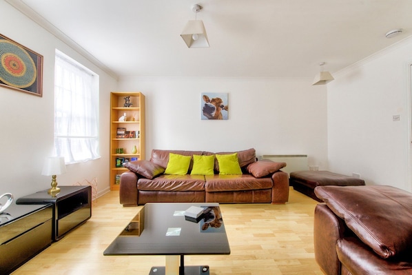 A living room with a brown leather sofa, a glass coffee table, a bookshelf, and a painting on the wall.