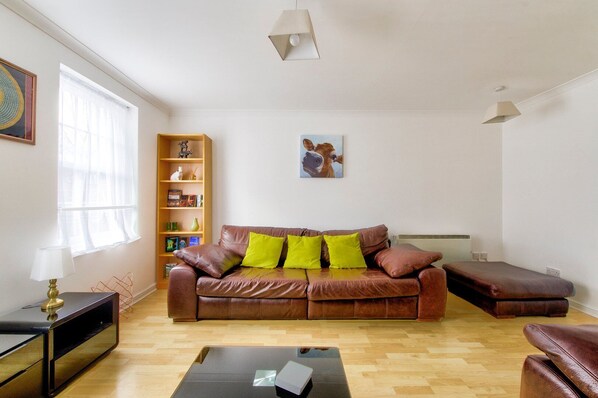 A living room with a brown leather sofa, a coffee table, a bookshelf, and a painting on the wall. - GuestReady - Spacious 2BR in Leith (Leith)